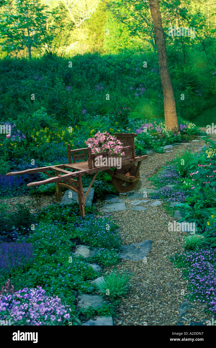 Mit Blumen gefüllte antike Schubkarre auf einem Schotterweg in einem idyllischen schattigen Garten entlang des Pathway, Missouri, USA Stockfoto