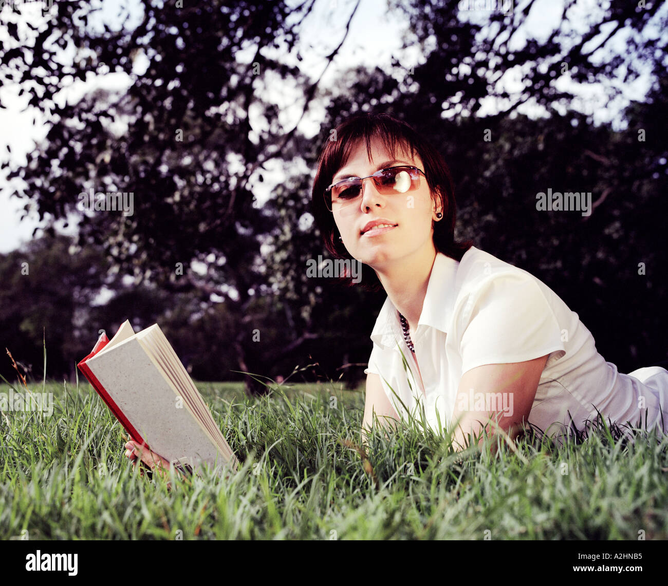 Eine junge 20 s 30 s Frauen tragen weiße Top Verlegung auf dem Rasen in einem Park, lesen ein Buch Bäume im Hintergrund Stockfoto