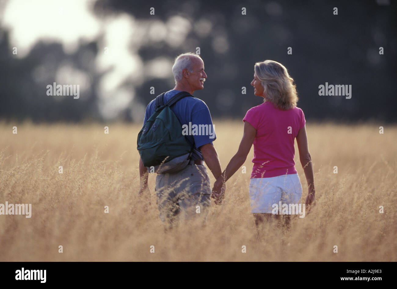 Lässig gekleidet älteres paar Lächeln einander an, wie sie hand in hand durch hip hohen gelben Gräser gehen Stockfoto