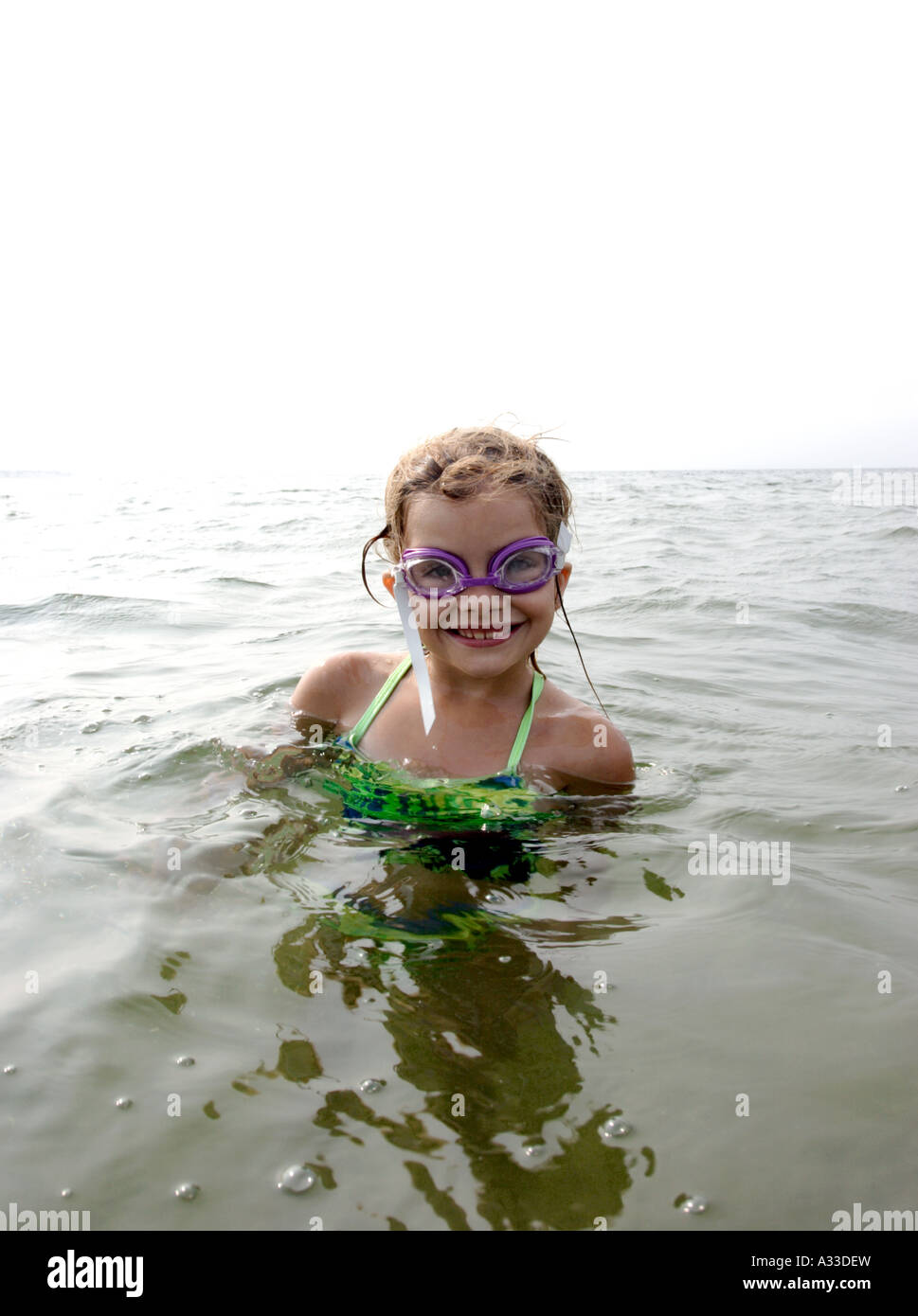 junges Mädchen mit sandigen blonde Haare mit grünen Badeanzug, lila Brillen in den Ozean lächelnd Stockfoto