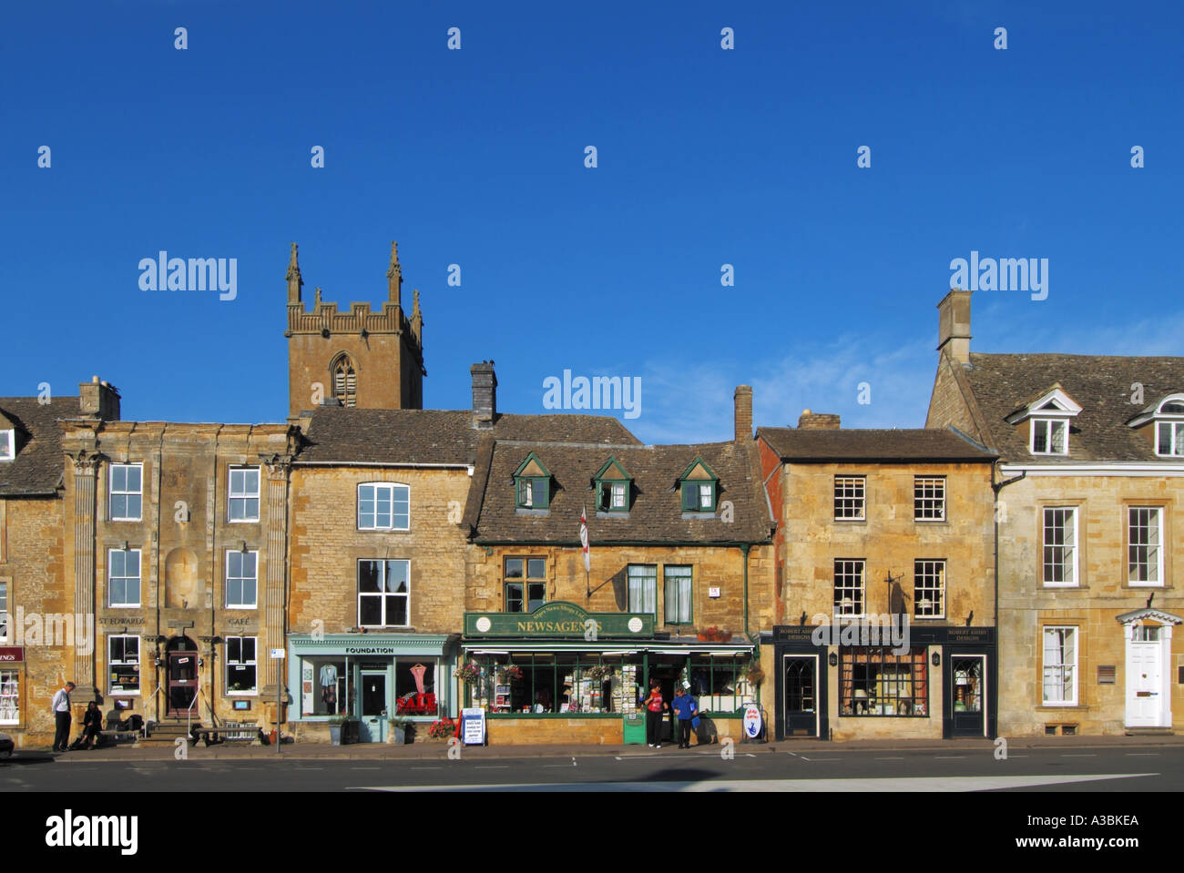 Erleben Sie den Tag des blauen Himmels der Wold in der Straßenszene der Stadt Cotswolds Market mit Kirchturm dahinter in einem Teil des Market Square Gloucestershire England Großbritannien Stockfoto
