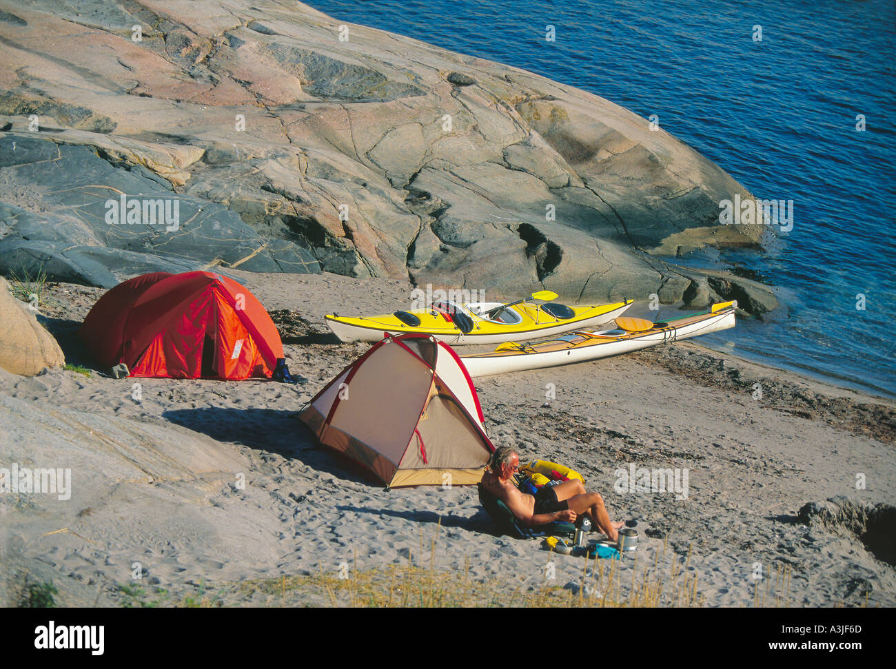 Ein Kajakfahrer genießen ruhige sonnige umgeben Morgen zwei Zelte und Seekajaks auf winzigen Sandstrand von Felsen Schweden Bohusland Stockfoto