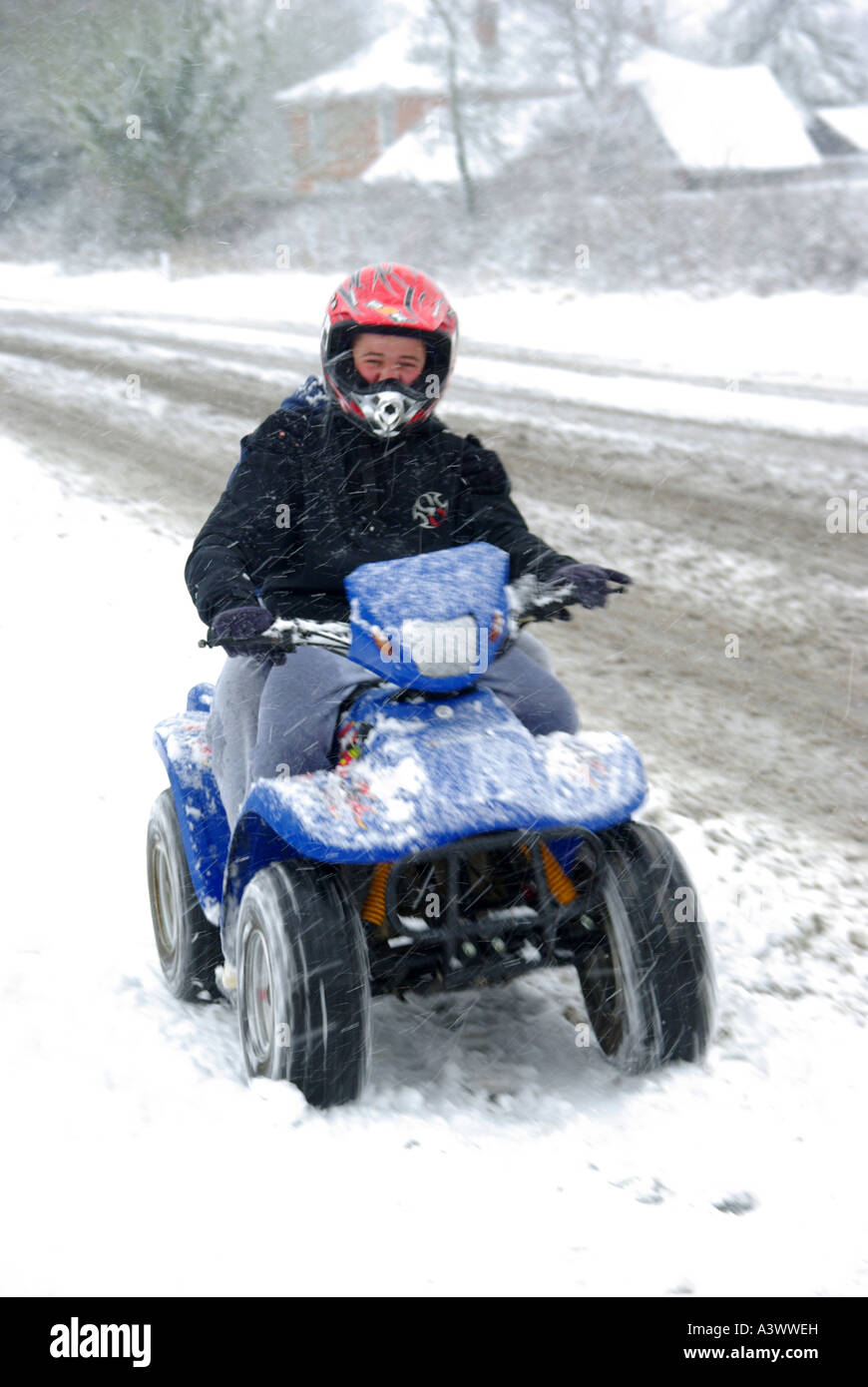 Schneefall in den jungen Menschen in roten Sturzhelm fahren quad bike schnell entlang verschneite Dorf Pflaster unklar, ob männlich oder weiblich Essex England Großbritannien Stockfoto