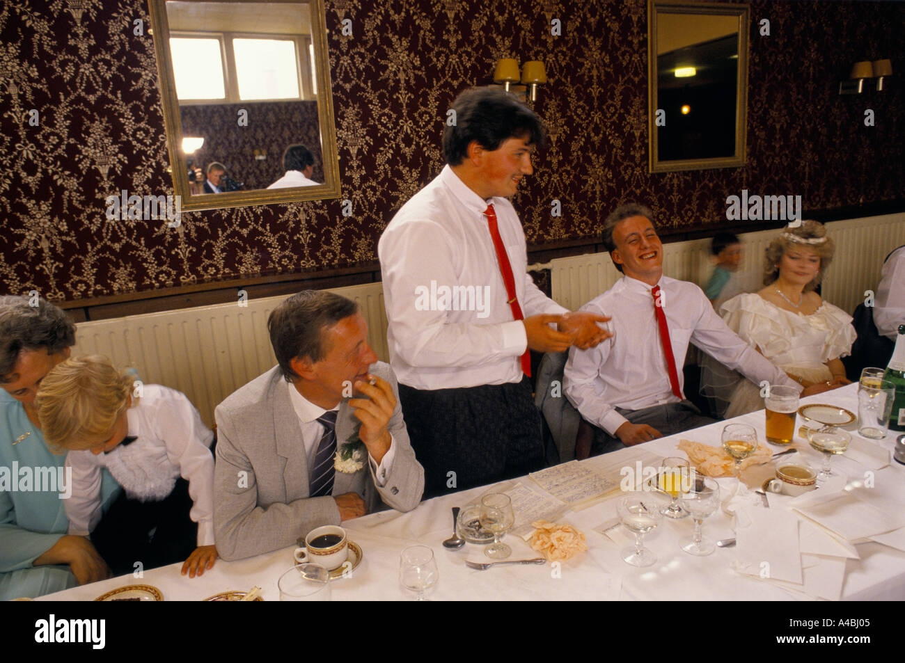 ein Mann mit einem dünnen Schnurrbart steht und hält eine Rede an einem langen Tisch bei einer Hochzeit der Bräutigam lacht die Braut ist nicht und andere Gäste, darunter ein kleines Kind suchen anderswo Stockfoto
