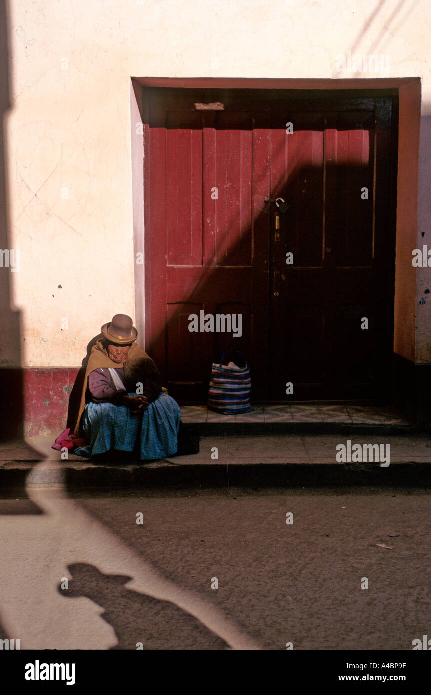 Paucartambo, Peru. Eine Quechua-Frau in traditioneller Kleidung vor einer verschlossenen Tür, halb im Schatten sitzen. Stockfoto