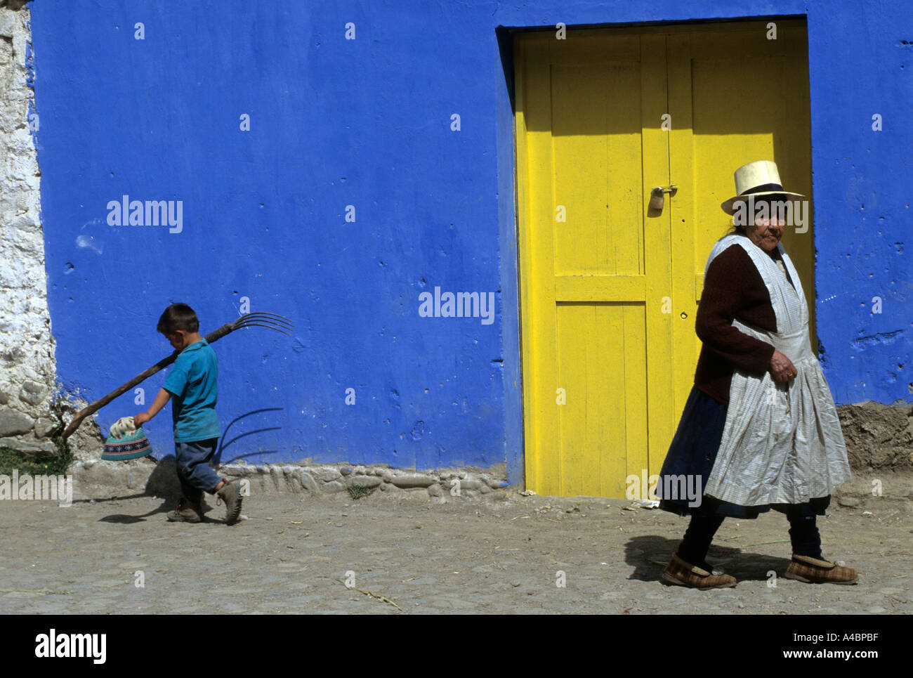 Ollantaytambo, Peru. Frau in Tracht und mit einer Gabel vor einer blauen Wand mit einer gelben Tür Boy. Stockfoto