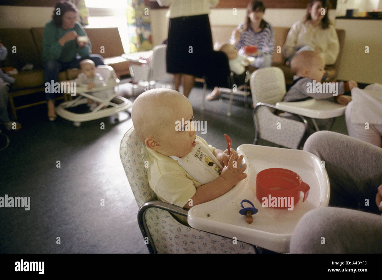 Ein Baby sitzt auf einem Stuhl vor seiner Mutter, einen roten Löffel umklammert. Stockfoto