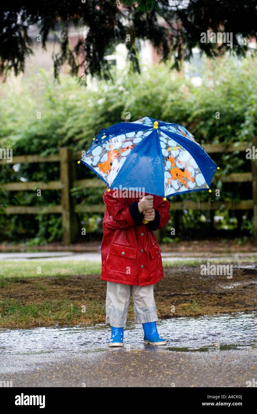 Kleiner Junge im roten Mantel und blaue Regenschirm plantschen in einer Pfütze in einem Park auf seine eigene bei Wetter Sturm. Stockfoto