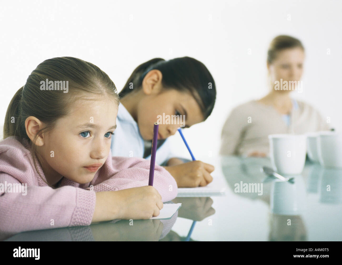 Zwei Mädchen sitzen am Tisch schreiben, Frau im Hintergrund Stockfoto