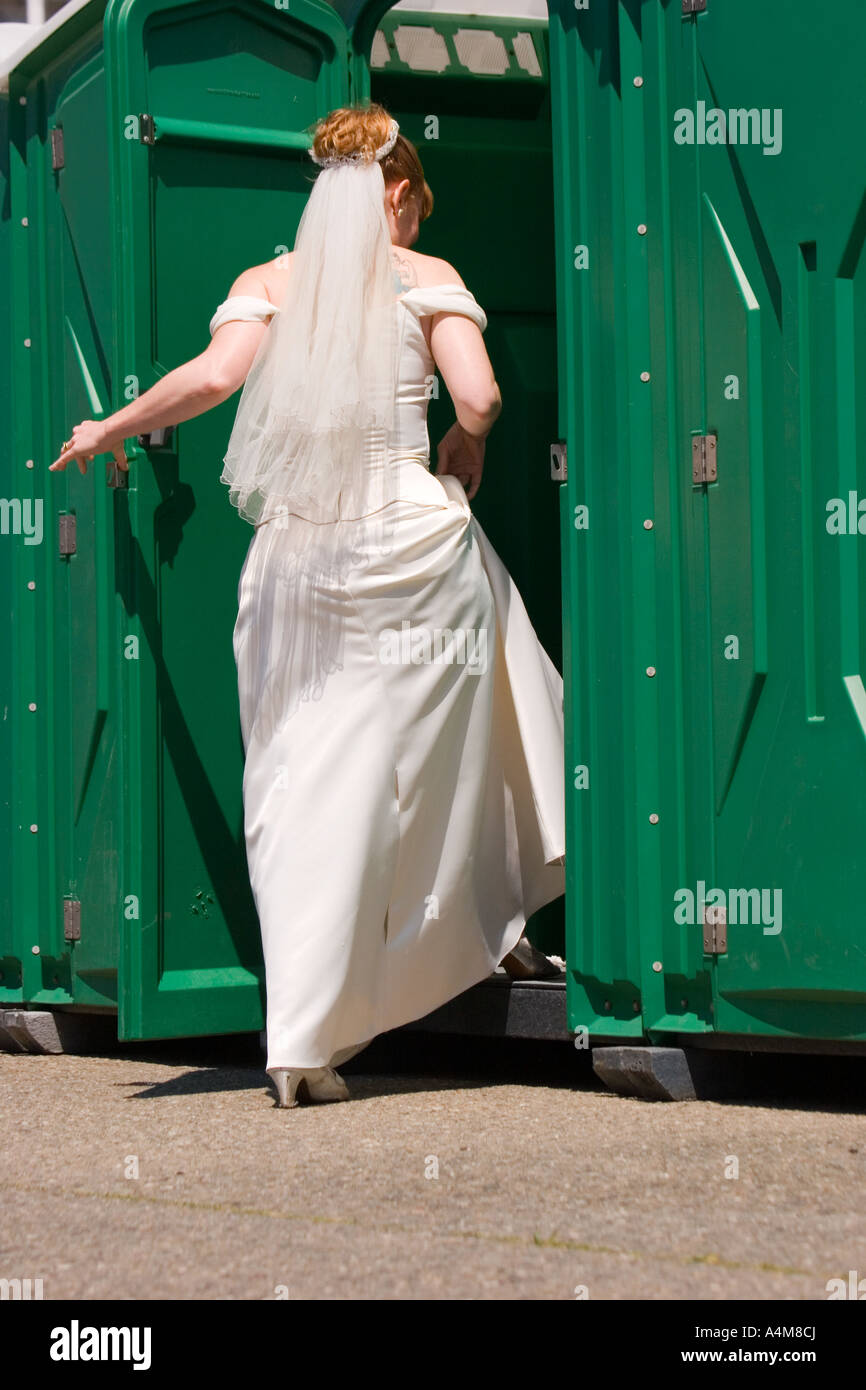 Eine Braut im weißen Kleid und Schleier betritt ein Port-a-Töpfchen. Stockfoto