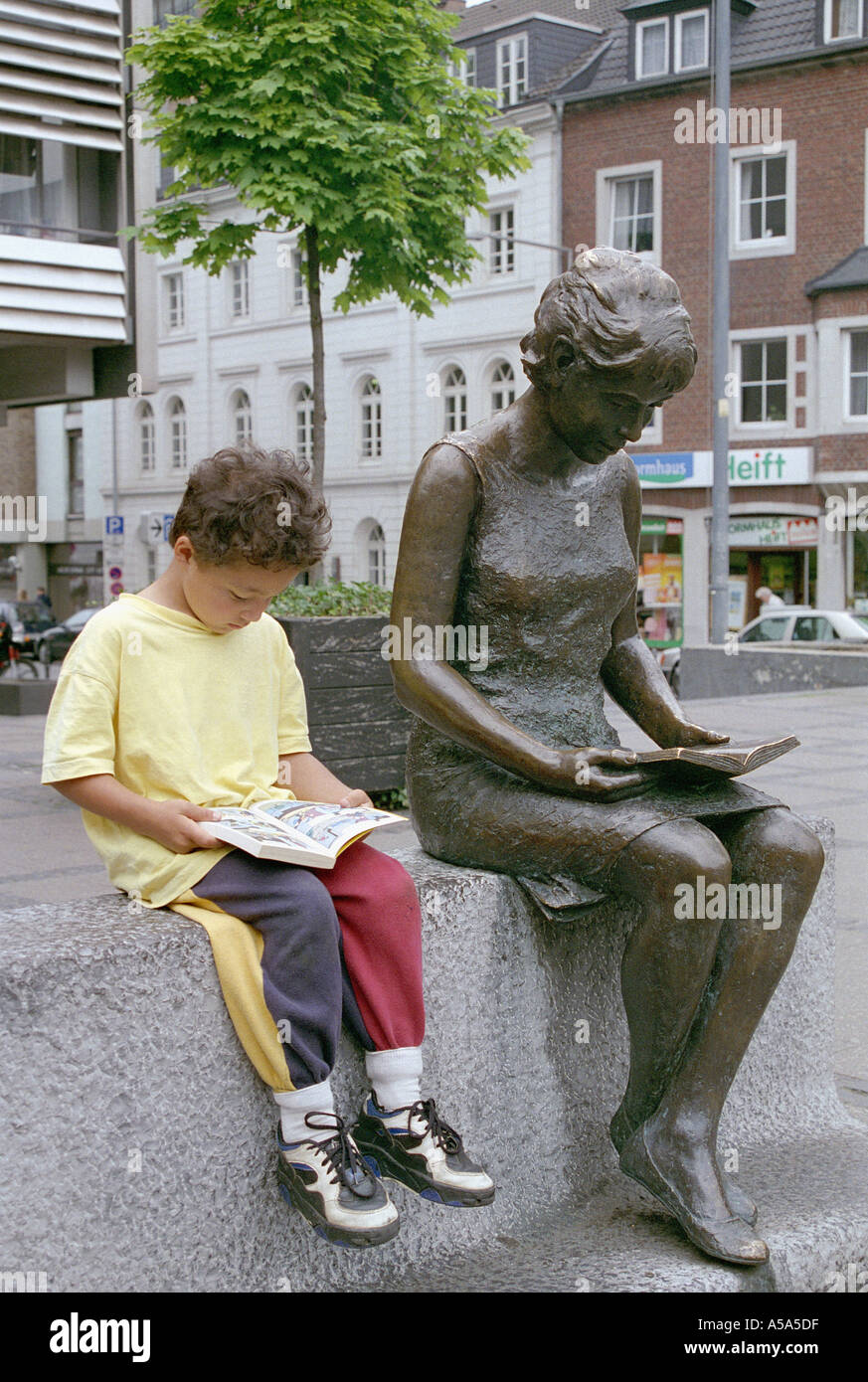 Ein kleiner Junge liest neben einer Statue von jemand, der liest, in Aachen, Deutschland. Stockfoto