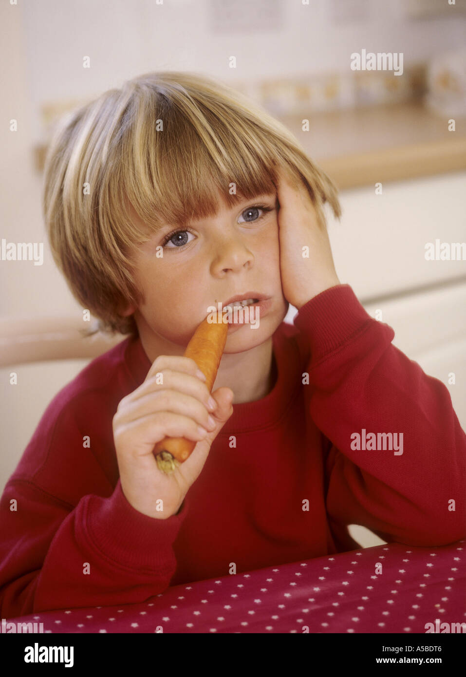Kleiner Junge sitzt in einer Küche, beißen in eine Möhre - unglücklich Stockfoto