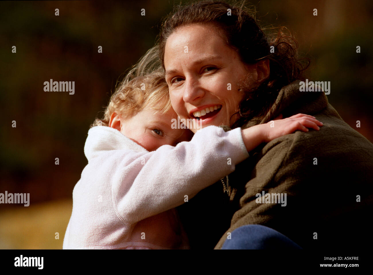 Eine Mutter bückt sich neben ihrer kleinen Tochter, wie das Mädchen ihre Mutter umarmt und sie auf den Betrachter Lächeln Stockfoto