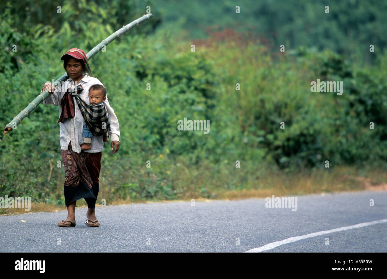 Mutter mit einem Bambusstab und tragen ihr Kind auf ihre Taille geht auf eine Asphaltstraße in Laos. Stockfoto