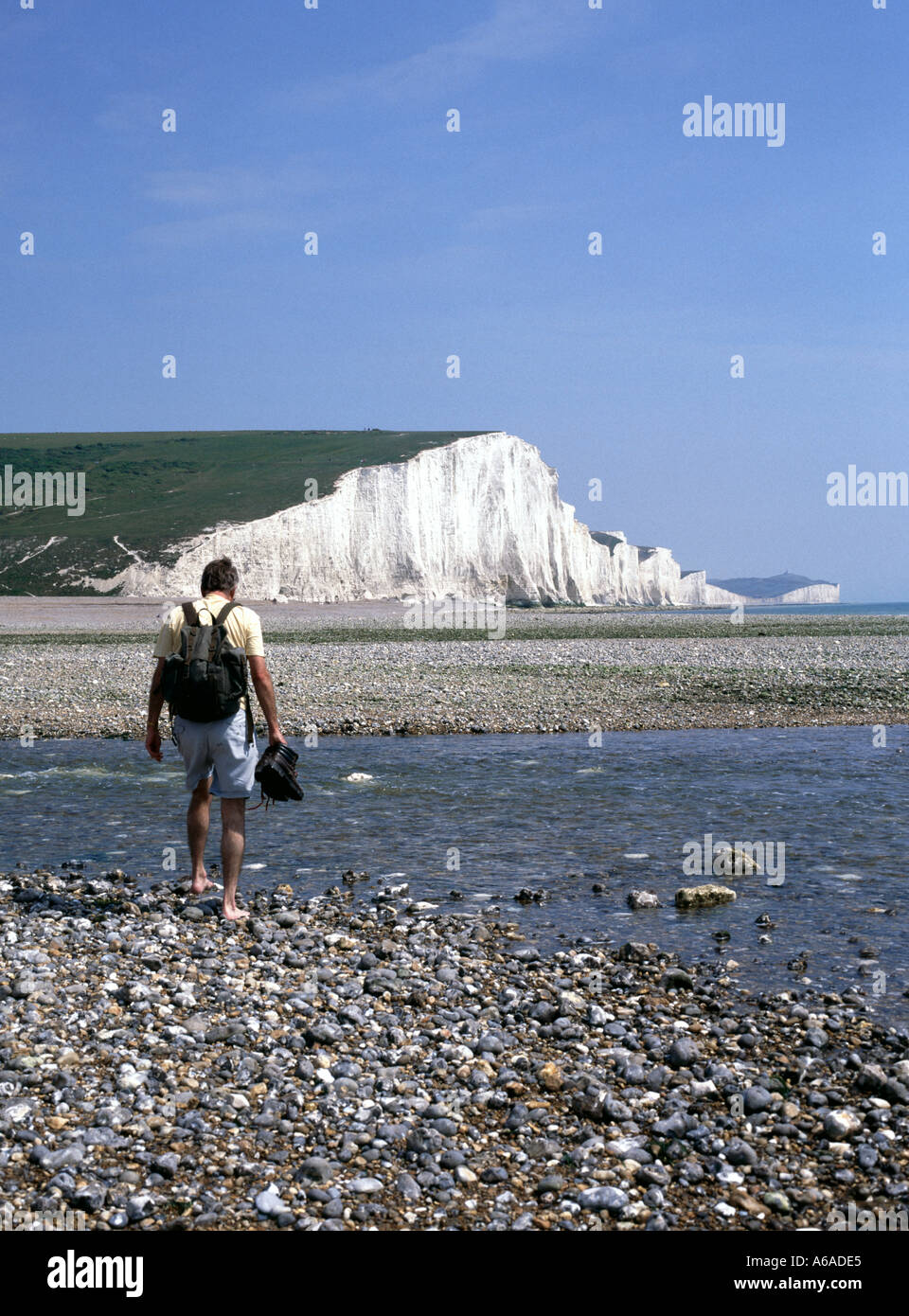Walker paddelt im Wasser über den Fluss Cuckmere Haven Mündung Mann Wanderschuhe und Rucksack Seven Sisters Kreidefelsen South Downs East Sussex UK Stockfoto