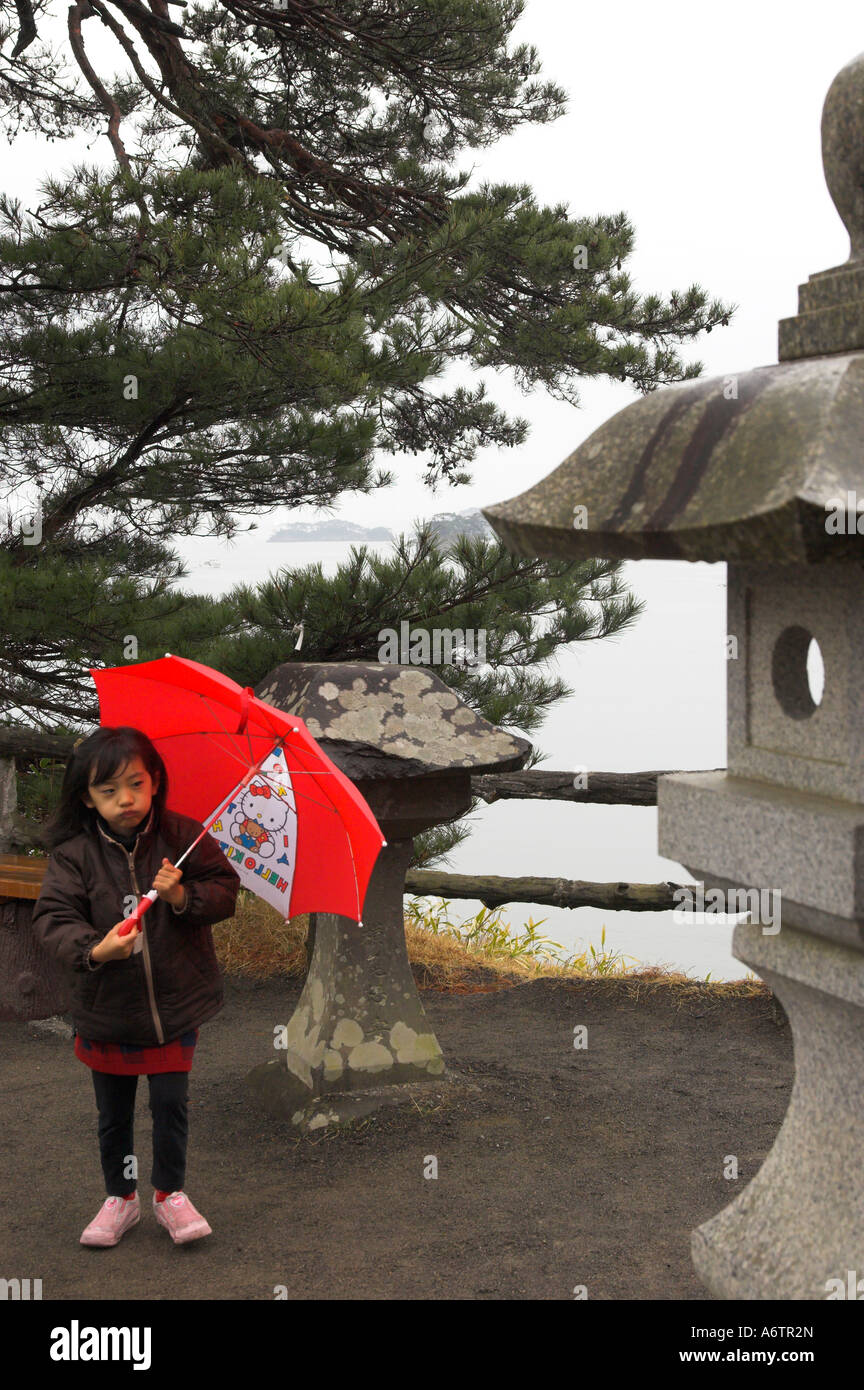 Nördlichen Honshu Miyagi in Japan ken Matsushima Bucht junge weibliche Kind hält einen roten Regenschirm an regnerischen Tag mit traditionellen Stein sta Stockfoto