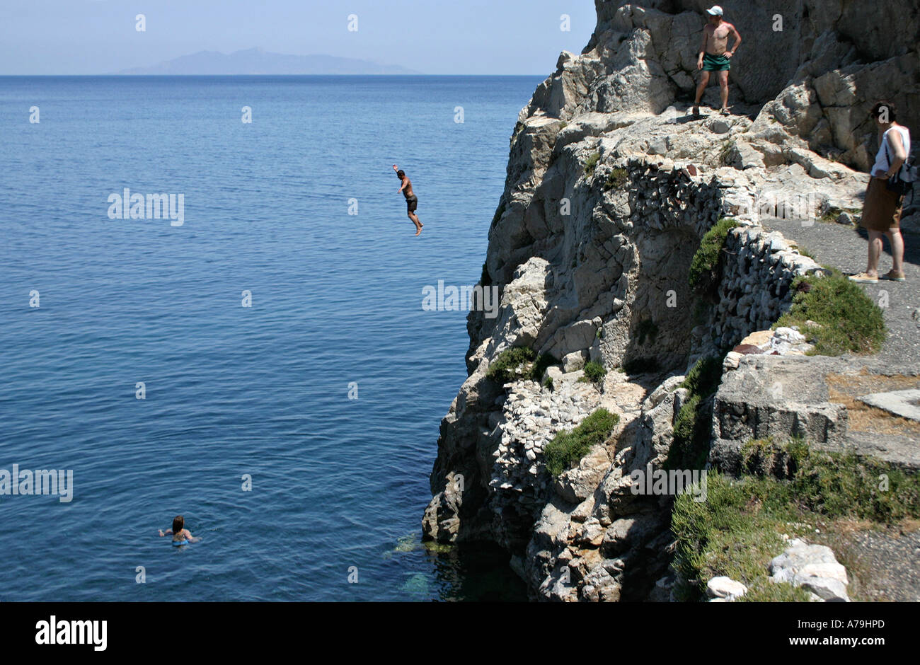 Erfrischender Sprung: Ein junger Mann von den Klippen springt ein Ende von Kamari Beach in die dunklen blauen Wasser ein Frau hat nur vorausgegangen Stockfoto