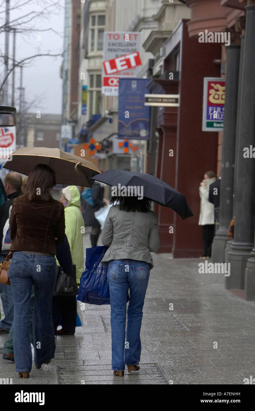 vertikale zwei junge Frauen tragen Regenschirme hinunter eine nasse Einkaufsstraße im Stadtzentrum zu Fuß entfernt von der Kamera Stockfoto