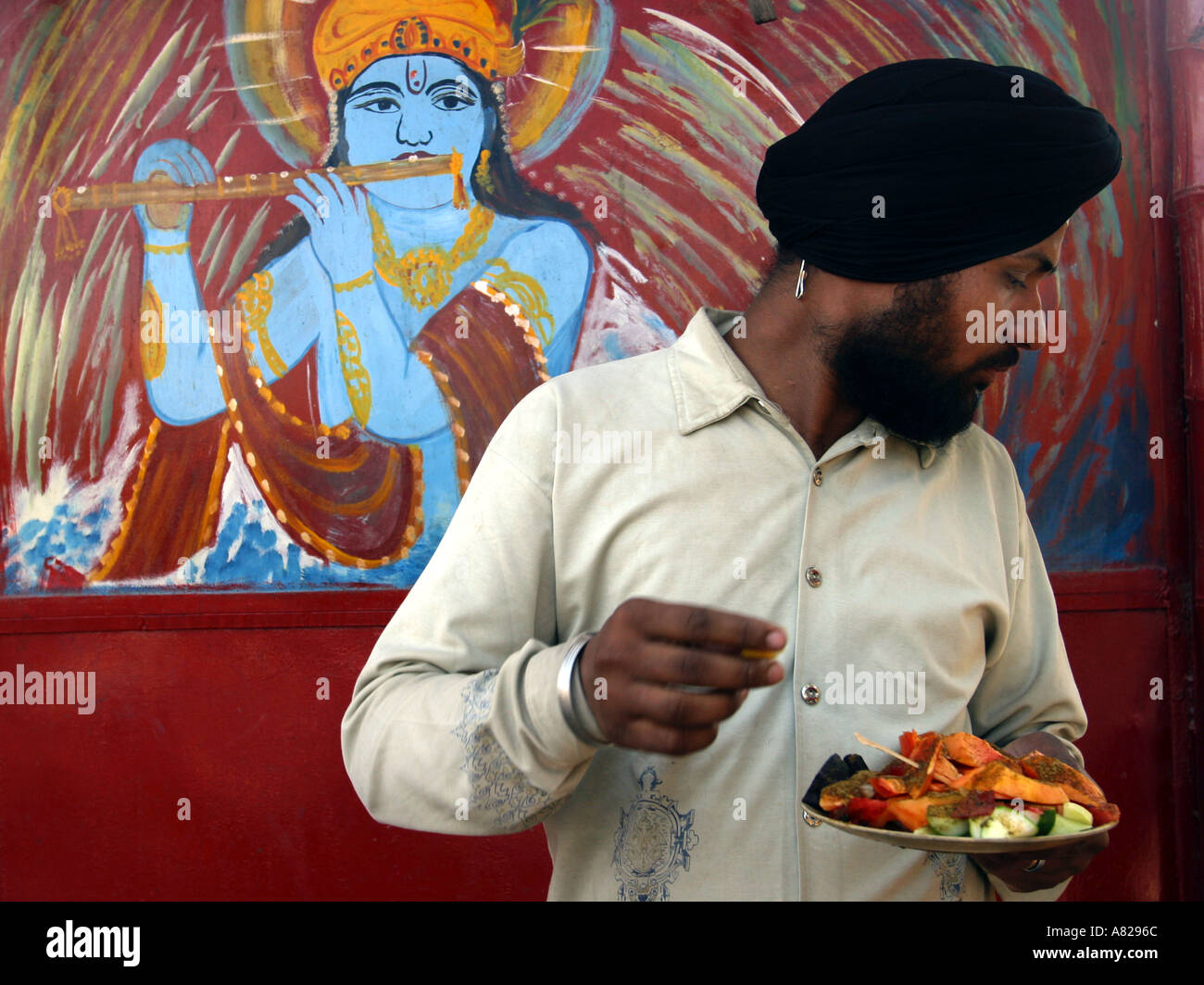 Ein Sikh Mann isst ein Essen A Gemälde Krishnas Flötenspiel im Hintergrund in Delhi Indien Stockfoto