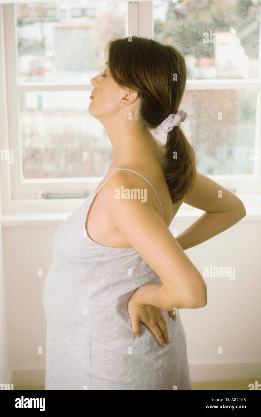 Hochschwangere Frau, die durch ein Fenster halten Sie ihren Bauch mit Rückenschmerzen Stockfoto