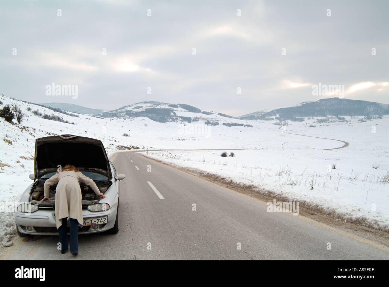 Junge Frau, die unter der Motorhaube ihres Autos kümmern, gestrandet auf einer abgelegenen Straße im Winter nach einer Pause nach unten Stockfoto