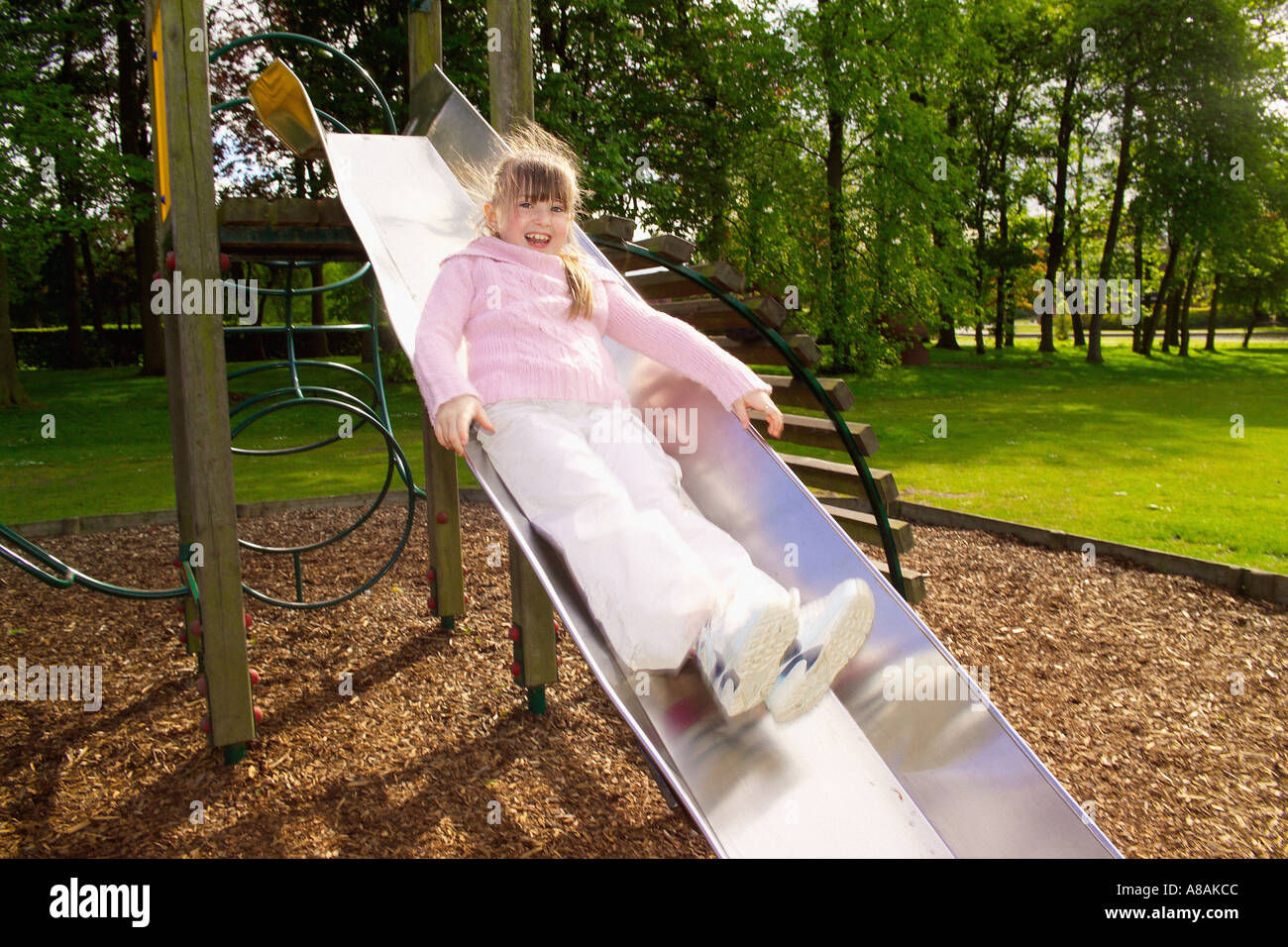 Ein junges Mädchen lacht, als sie an einem kalten, aber sonnigen Sommertag auf eine Edelstahlrutsche auf die weichen Rindenabfälle im Rowntree Park gleitet. York. Stockfoto