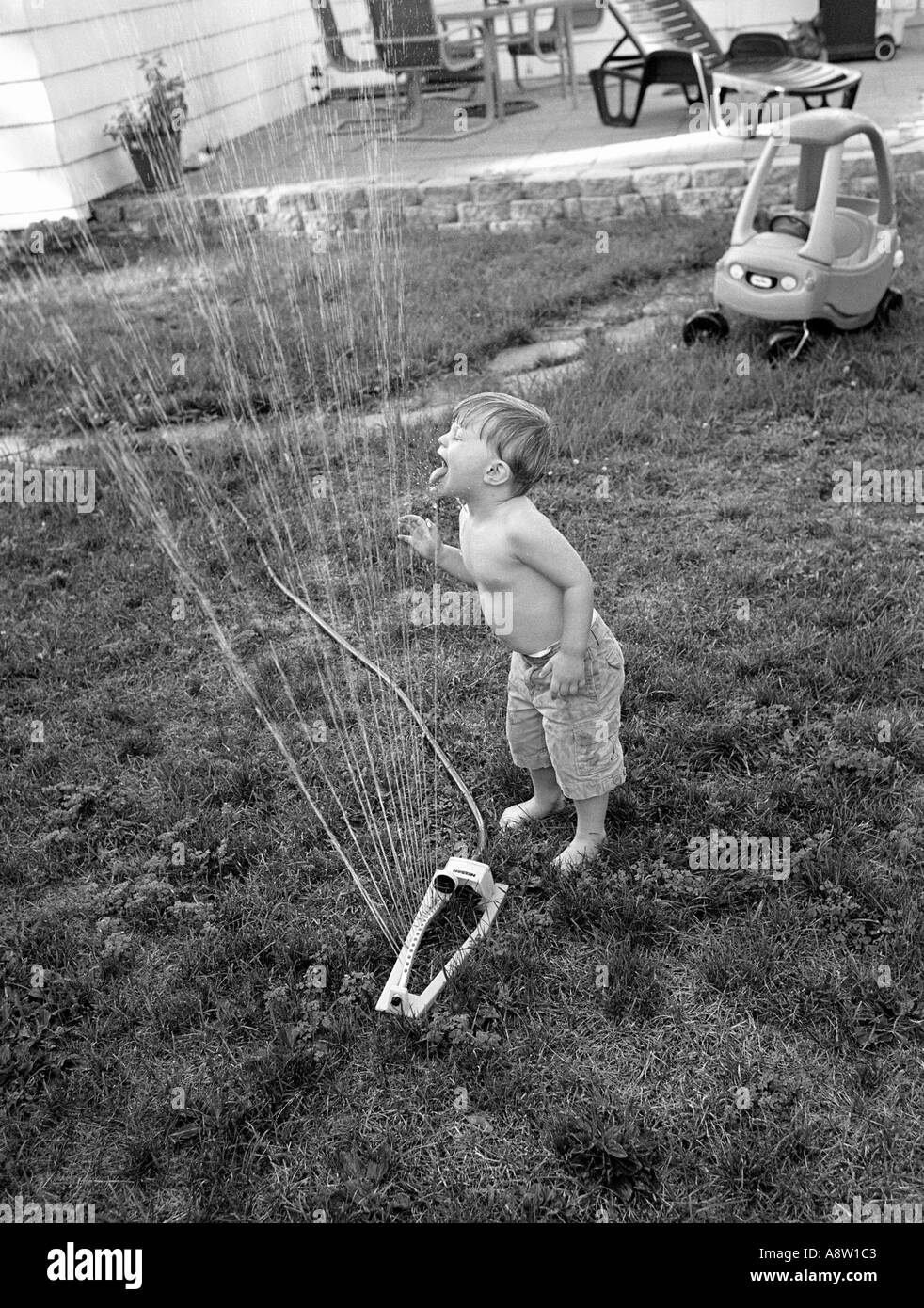 Kleiner Junge spielt mit Rasen-Sprinkler-Trinkwasser und Abkühlung an einem heißen Sommertag in seinem Hinterhof Stockfoto