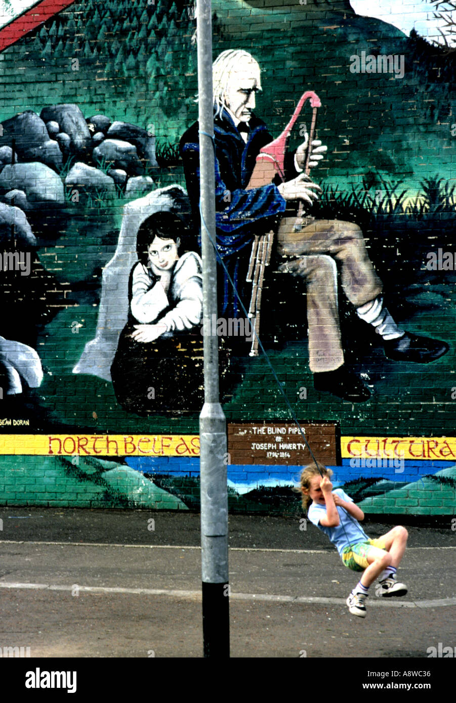 Ein Kind schwingt von einem Laternenpfahl gegen ein Wandbild Backgrond von einem blinden Piper in Belfast N Irland Stockfoto