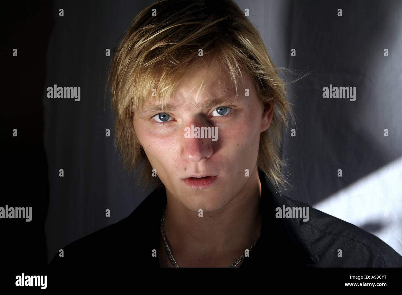 Nahaufnahme eines jungen Mannes mit blonden Haaren und intensiven blauen Augen, der direkt in die Kamera blickt, mit dramatischer Beleuchtung. Stockfoto