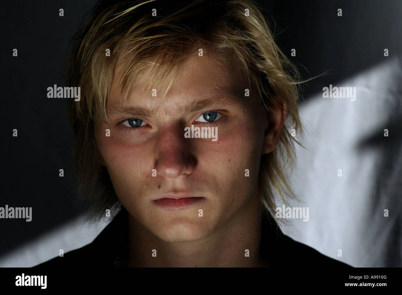 Nahaufnahme eines jungen Mannes mit blonden Haaren und intensiven blauen Augen, der direkt in die Kamera blickt, mit dramatischer Beleuchtung. Stockfoto