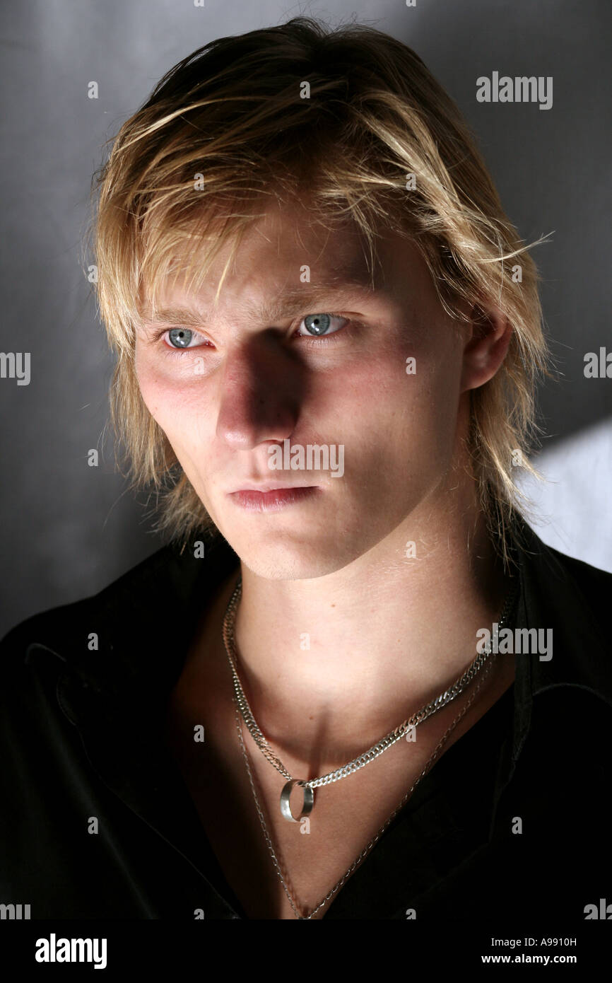 Porträt eines jungen Mannes mit blondem Haar, einer Kette und intensiven blauen Augen, hervorgehoben durch dramatische Beleuchtung und Schatten im Studio. Stockfoto