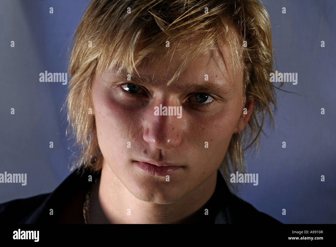 Nahaufnahme eines jungen Mannes mit blonden Haaren und intensiven blauen Augen, der direkt in die Kamera blickt, mit dramatischer Beleuchtung. Stockfoto