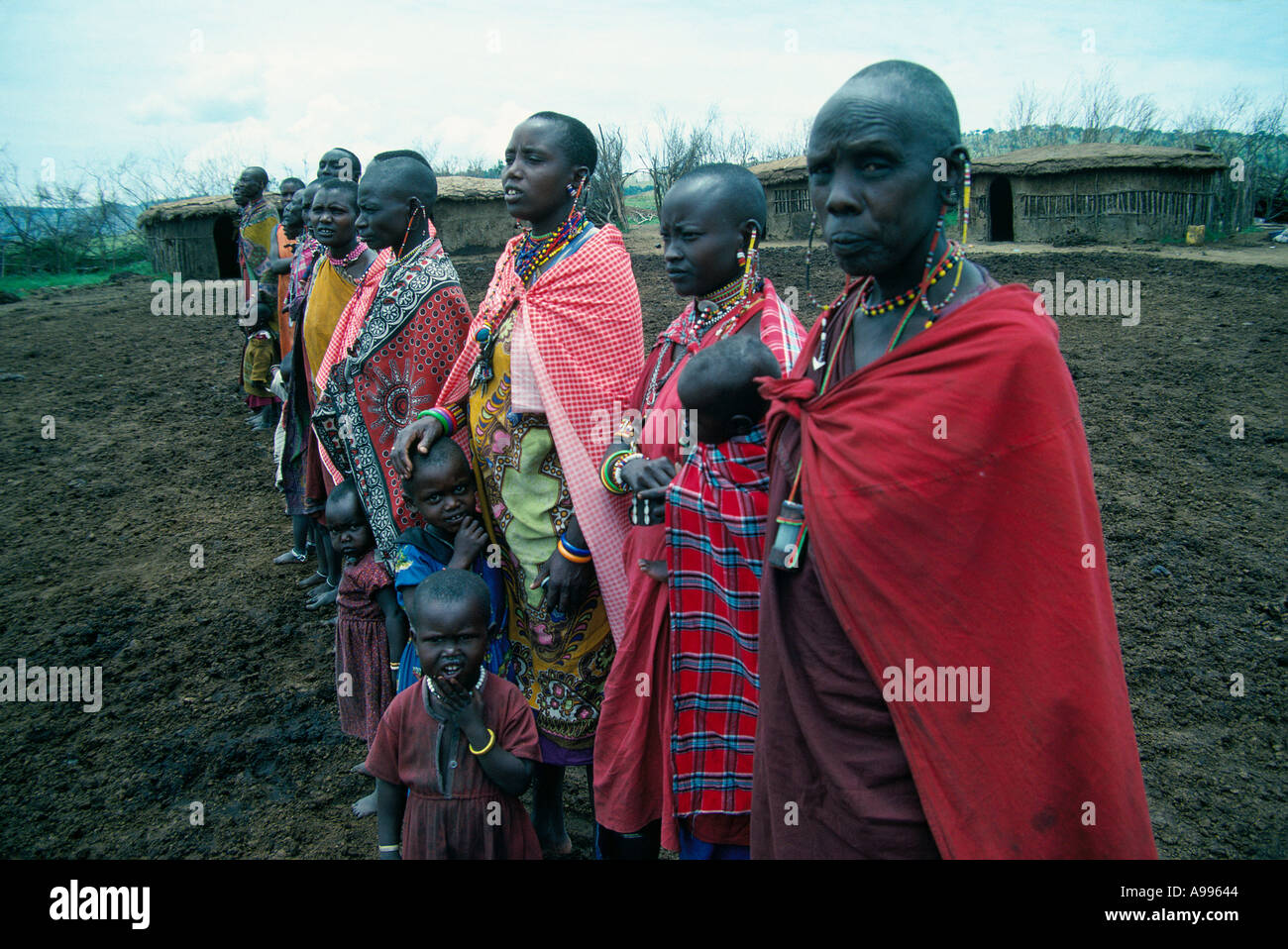 Masai Frauen und Kinder in bunten Gewändern stehen im Mittelpunkt ihres Dorfes in der Nähe ihrer Hütten Masai Mara Wildreservat Kenia Stockfoto