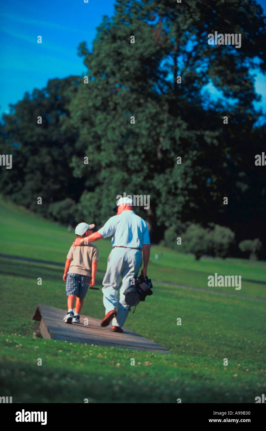 Ein Vater mit seinem Golf-Bag und Clubs hat seinen Arm um seine Schultern Sohn, wie sie über eine Brücke auf einem Golfplatz gehen Stockfoto