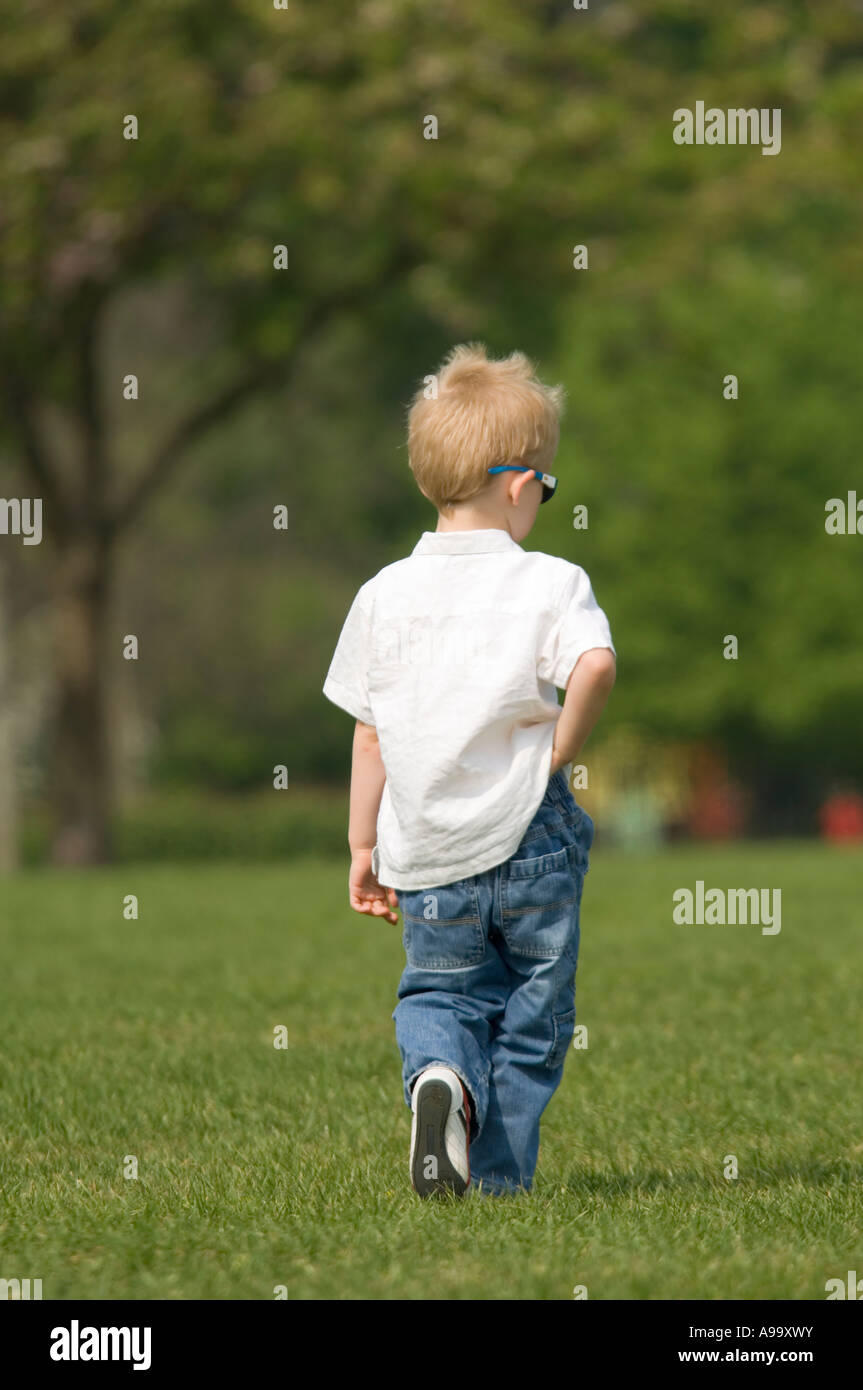 Eine blonde junge trägt ein weißes Hemd, Jeans und Sonne Brille in den Park zu Fuß entfernt mit dem Rücken zur Kamera an einem sonnigen Tag. Stockfoto