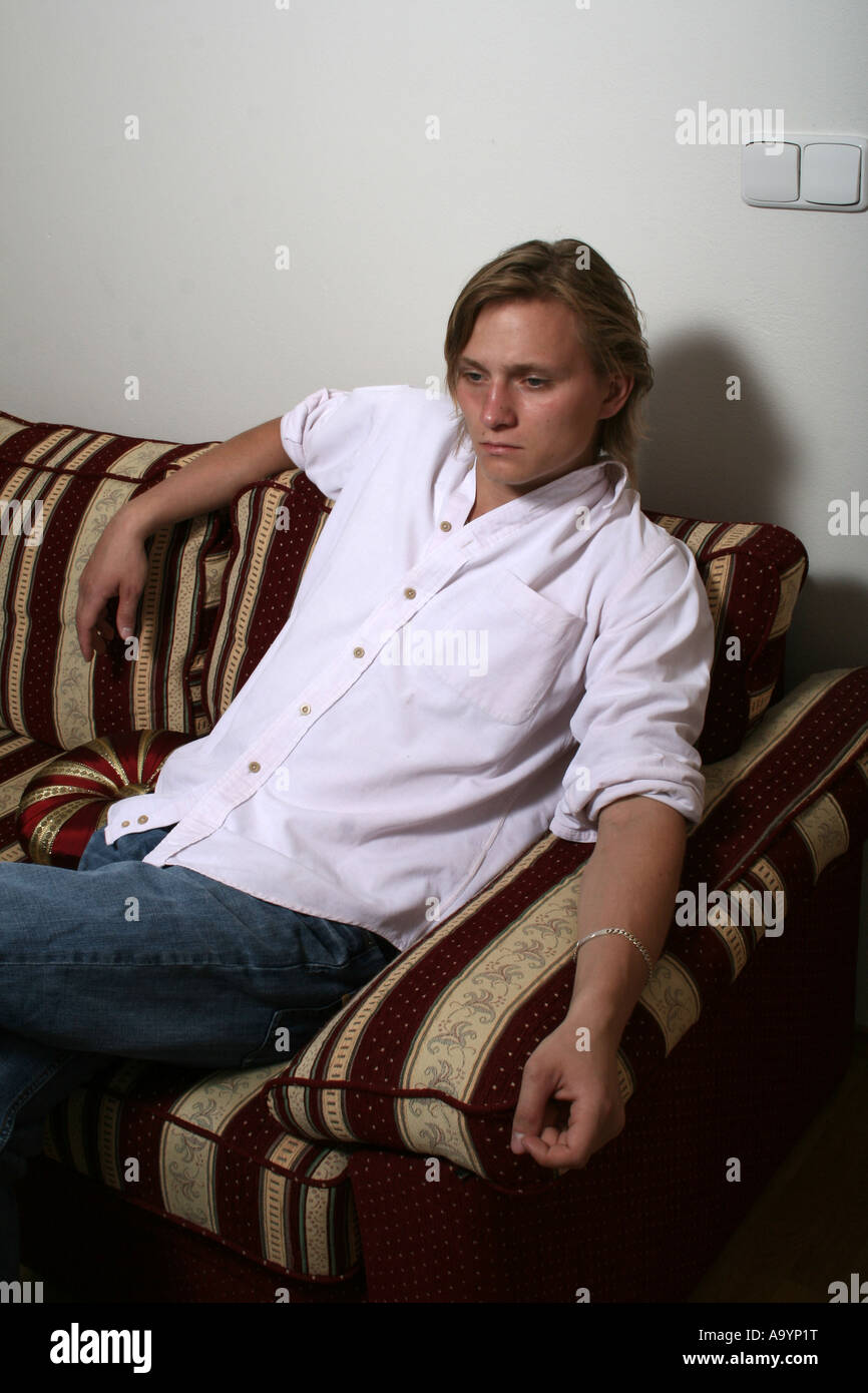 Entspannter junger Mann in lässigem weißem Hemd, der auf einem gemusterten Sofa sitzt, tief in Gedanken, vor einem schlichten Hintergrund. Stockfoto