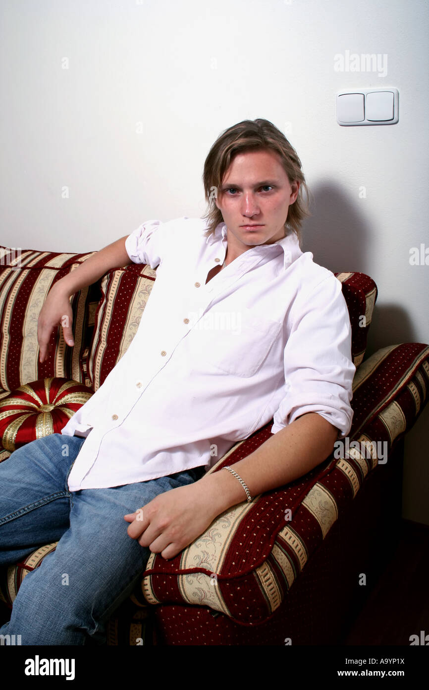 Selbstbewusster junger Mann in einem lässigen weißen Hemd, der auf einem gemusterten Sofa sitzt und sich mit einem entspannten und stilvollen Auftreten vor einer schlichten Wand posiert. Stockfoto