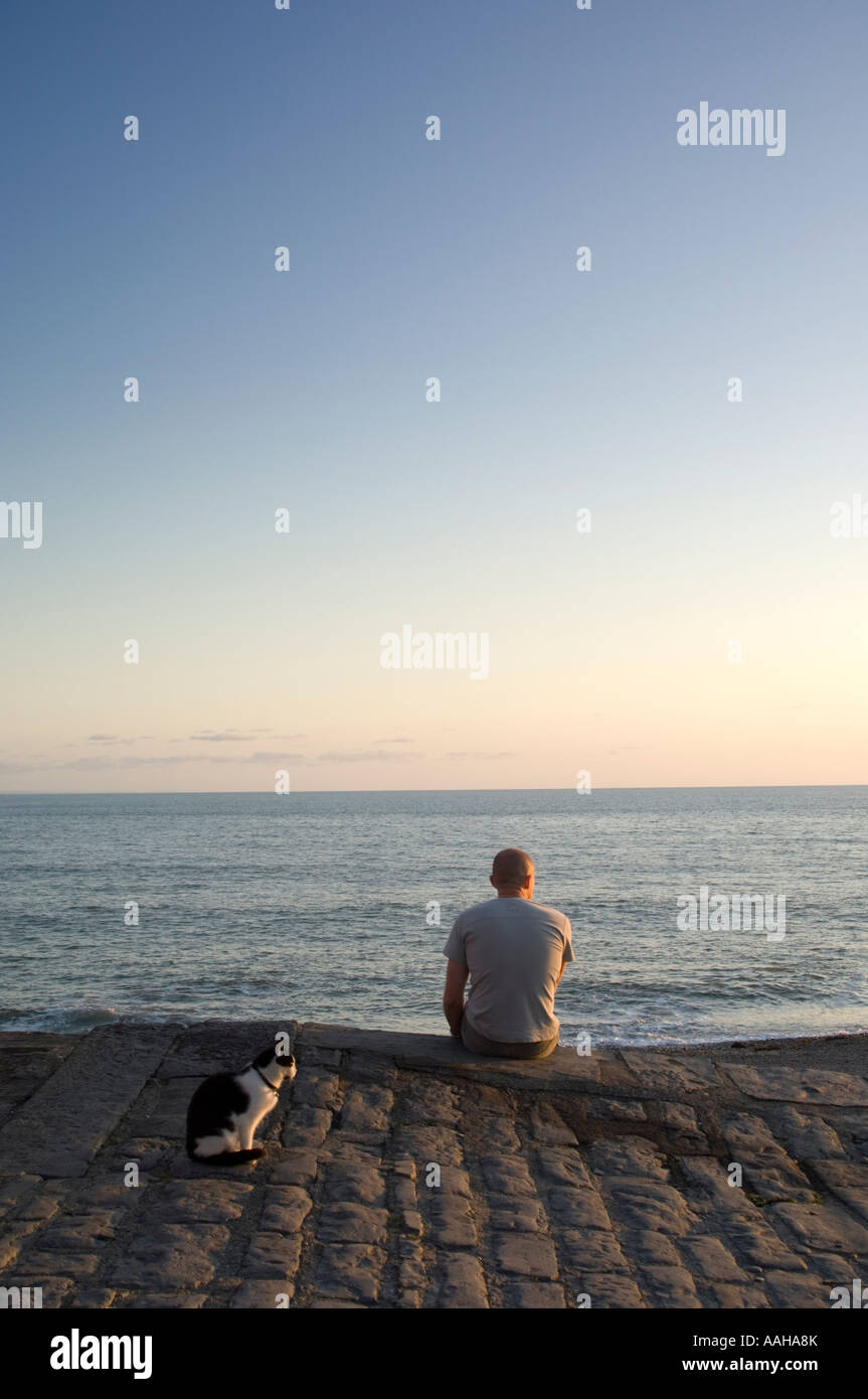 Mann sitzt auf Promenade Ufermauer Blick auf das Meer in einer ruhigen, friedlichen Sommerabend mit Katze an seiner Seite Aberystwyth, Wales Stockfoto