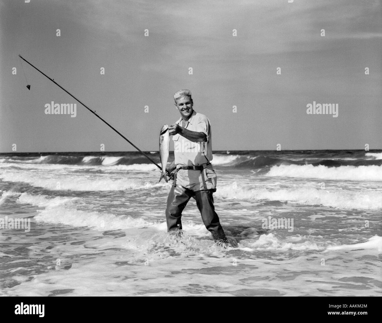 1950ER JAHRE ÄLTEREN MANN IN SURF IN WATVÖGEL, FISCH IN EINER HAND ANGELRUTE IN ANDEREN HÄLT Stockfoto
