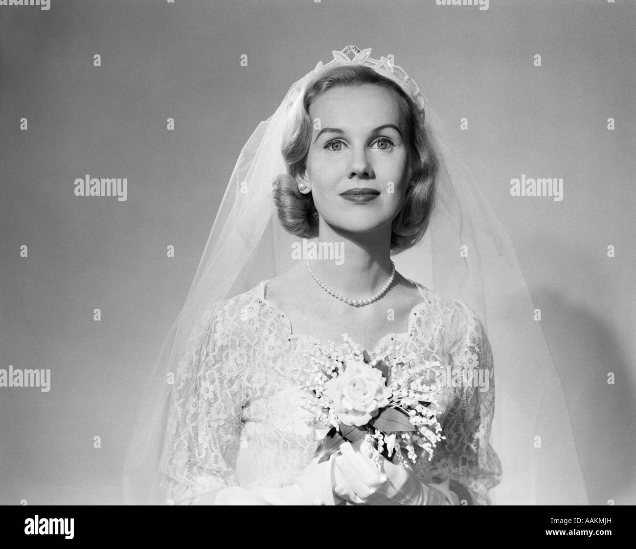 1950ER JAHRE PORTRAIT FRAU BRAUT TRAGEN WEIßE HOCHZEIT KLEID SCHLEIER PERLENSCHNUR HÄLT EINEN BLUMENSTRAUß BLICK IN DIE KAMERA Stockfoto