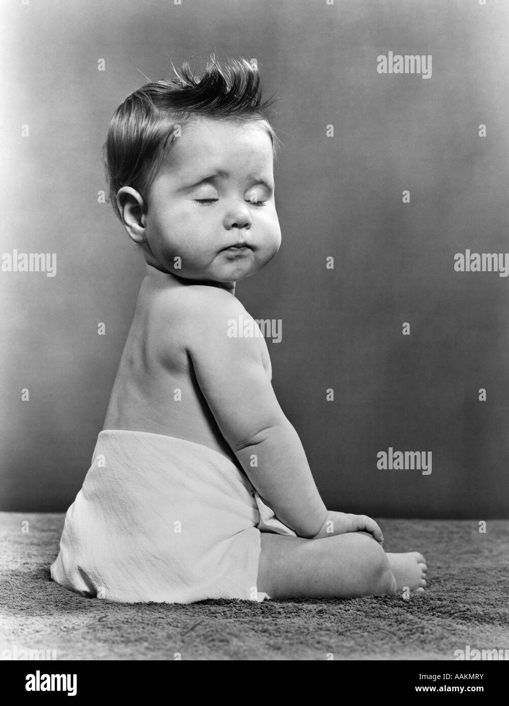 1940S 1950S BABY MIT ZURÜCK ZU DREHEN KAMERAKOPF BETRACHTER MIT GESCHLOSSENEN AUGEN SITZEN Stockfoto