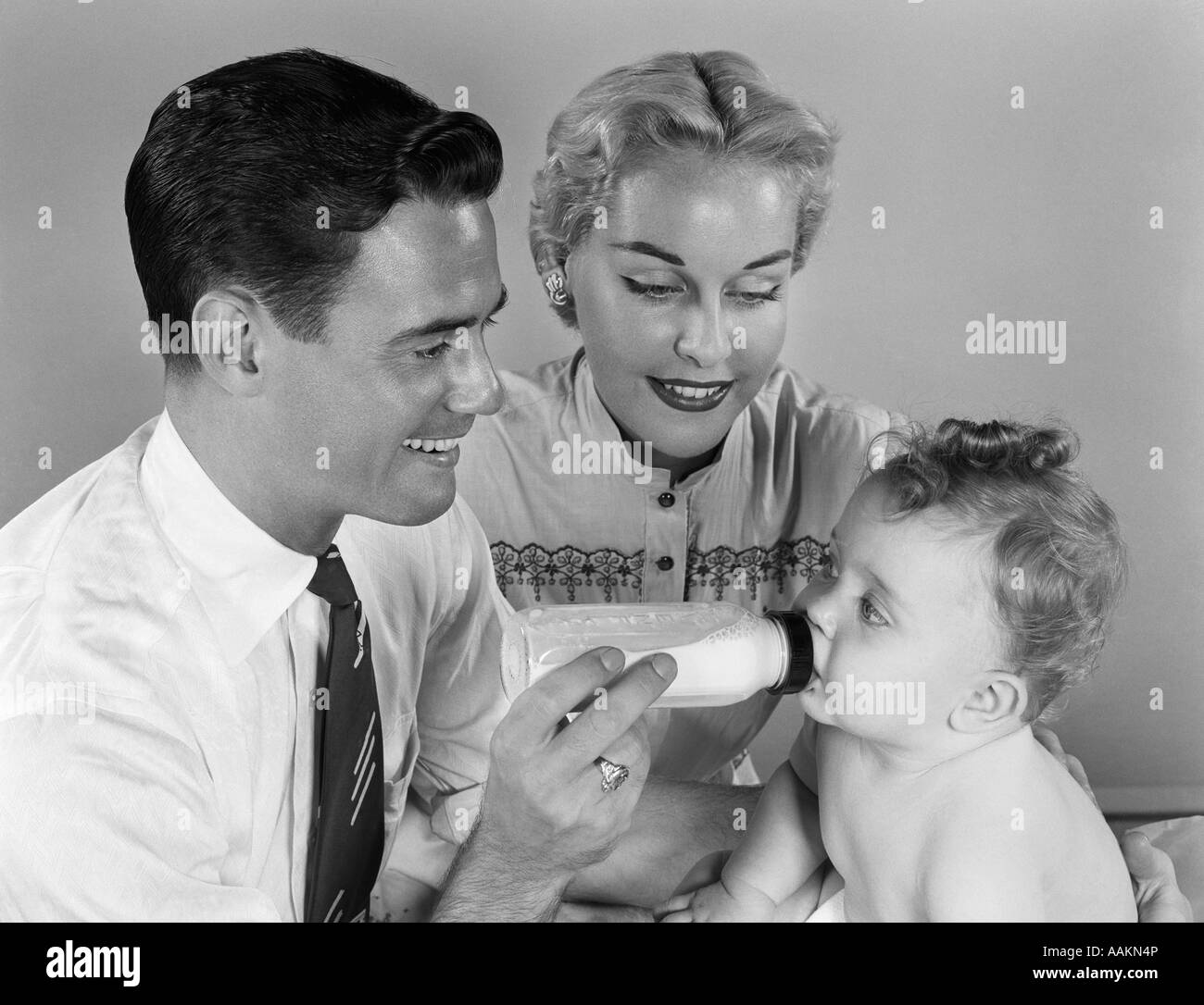 1950ER JAHREN LÄCHELND VATER FÜTTERUNG BABYMILCH AUS DER FLASCHE MIT MUTTER AT SEITE Stockfoto