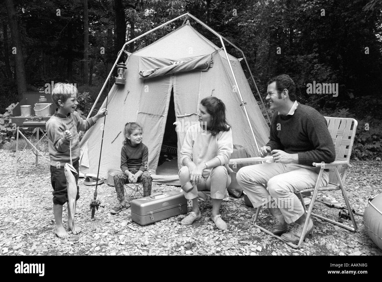 1970ER JAHREN VIERKÖPFIGE FAMILIE SITZT VOR DEM ZELT IM WALD SOHN MIT ANGELRUTE IN DER HAND & BIG FISH IN ANDEREN Stockfoto