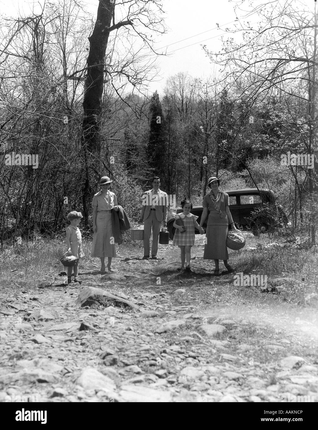 1930ER JAHRE FAMILIE ZU FUß ENTFERNT VON AUTO GEPARKT IN WÄLDERN MIT PICKNICK-KÖRBE & THERMOS-KANNEN Stockfoto