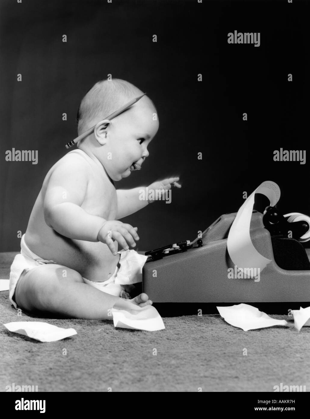 1960ER JAHREN SEITENANSICHT DES MOLLIG BABY SITZT HINTER RECHENMASCHINE MIT BLEISTIFT HINTER DEM OHR & BAND AUF BODEN VERSTREUT Stockfoto