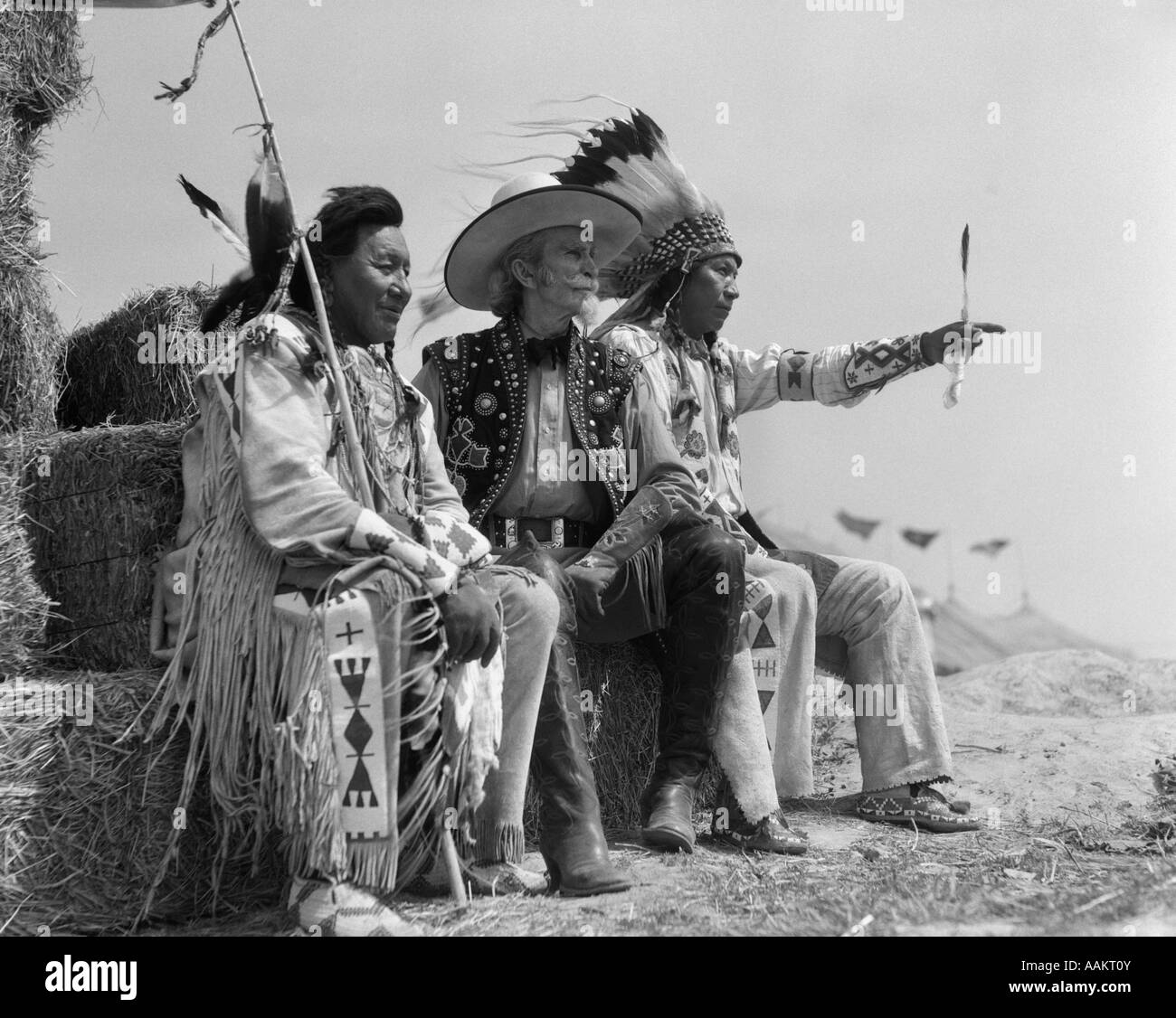 1940ER JAHREN PAAR DER INDIANER IN VOLLER TRACHT AUF STROHBALLEN SITZEND MIT COWBOY ZWISCHEN IHNEN Stockfoto