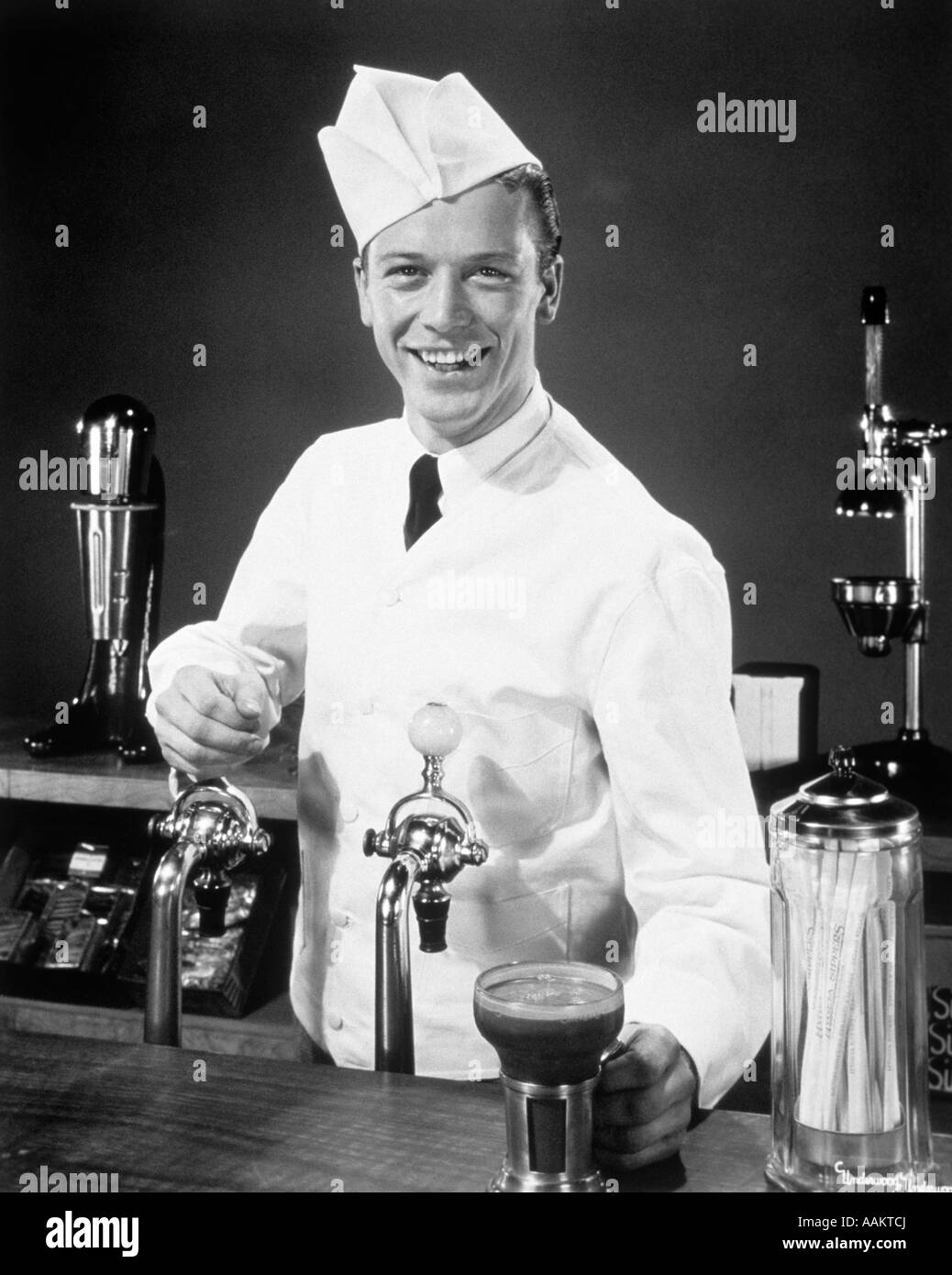 1940S 1950S LÄCHELNDE JUNGE MANN SODA JERK AM SCHALTER DIENEN EIN ICE CREAM SODA BLICK IN DIE KAMERA Stockfoto