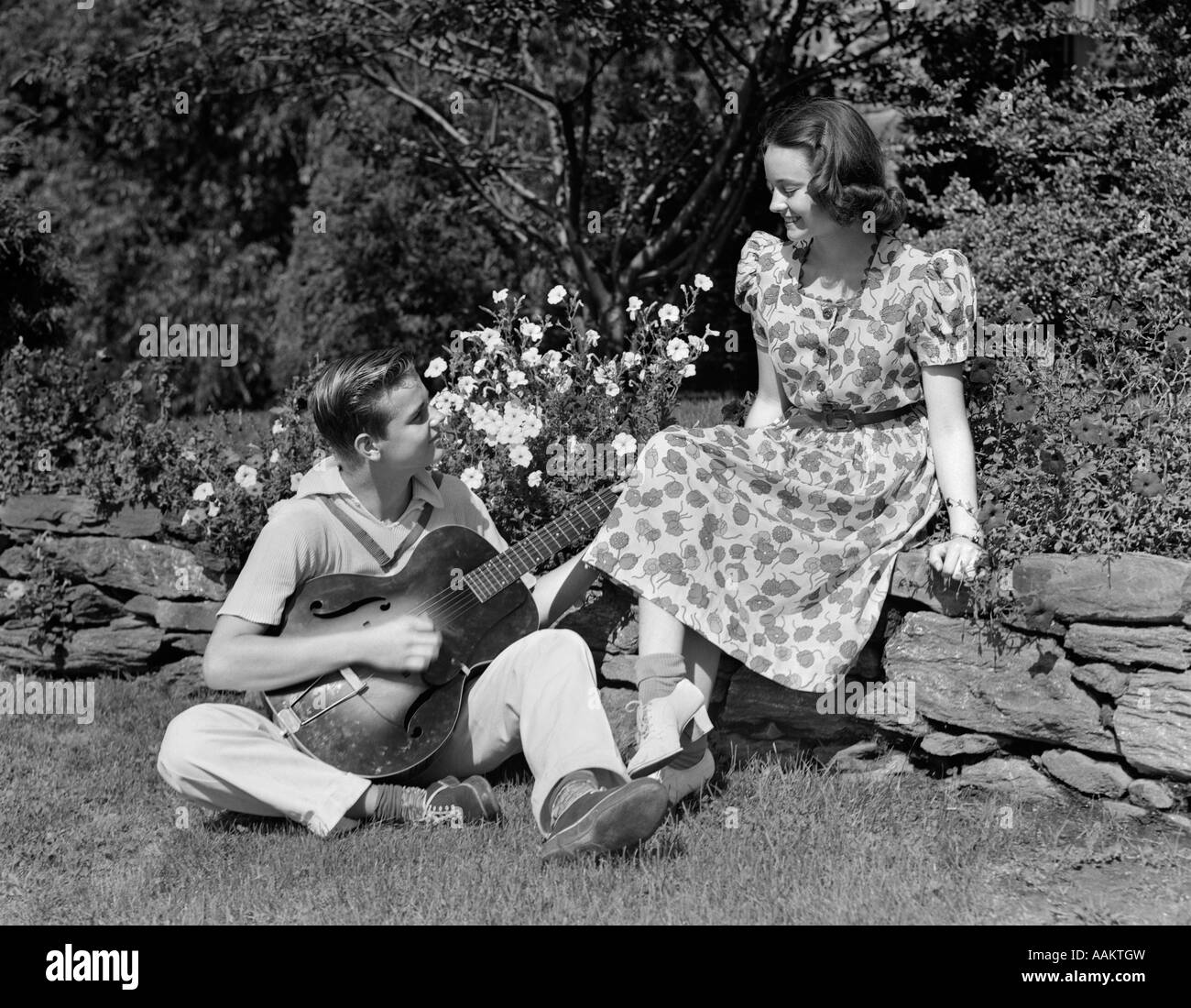 1940ER JAHRE JUNGES TEEN PAAR JUNGE SITZT IM RASEN SPIELEN GITARRE STÄNDCHEN MÄDCHEN SITZEN AUF STEINZAUN Stockfoto