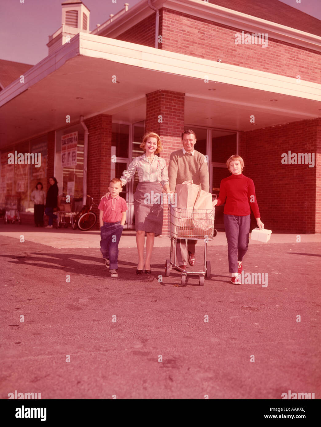 1960ER JAHRE FAMILIE SCHIEBEN EINKAUFSWAGEN VERLASSEN SUPERMARKT BLICK IN DIE KAMERA Stockfoto