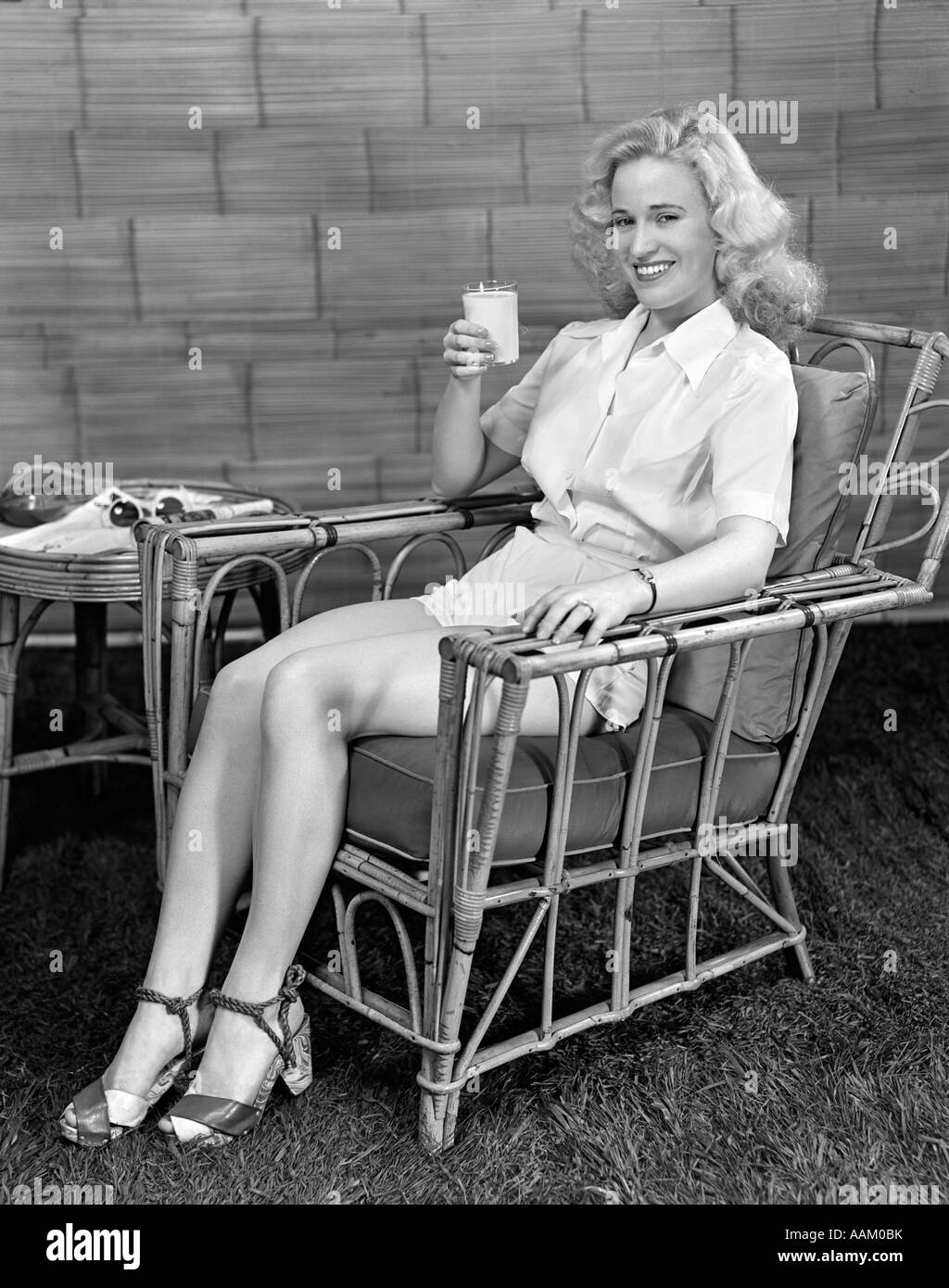 1940ER JAHRE BLONDE FRAU IN BAMBUS STUHL HÄLT GLAS MILCH TRAGEN WEIßE HOSE, BLICK IN DIE KAMERA LÄCHELN Stockfoto